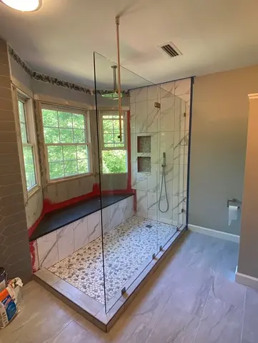 A walk-in shower with white and gray tile and a frameless glass splash panel in front of a bay window from Glass Doctor of Raleigh